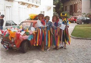 Grupo de pessoas vestidas com fatos coloridos e fitas, em frente a um carro decorado, numa rua de calçada durante uma festa ou desfile.
